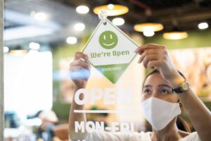 a female restaurant worker wearing a mask is flipping over the restaurant's sign to let customers know the restaurant is open