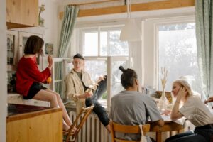 A group of four friends sit in a kitchen, on counters, and in the window as they talk and catch up.