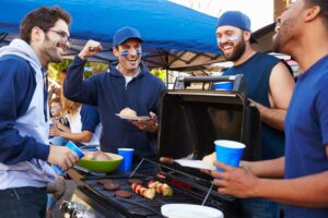 A group of friends stands around a grill at a sports tailgate while cooking food and talking