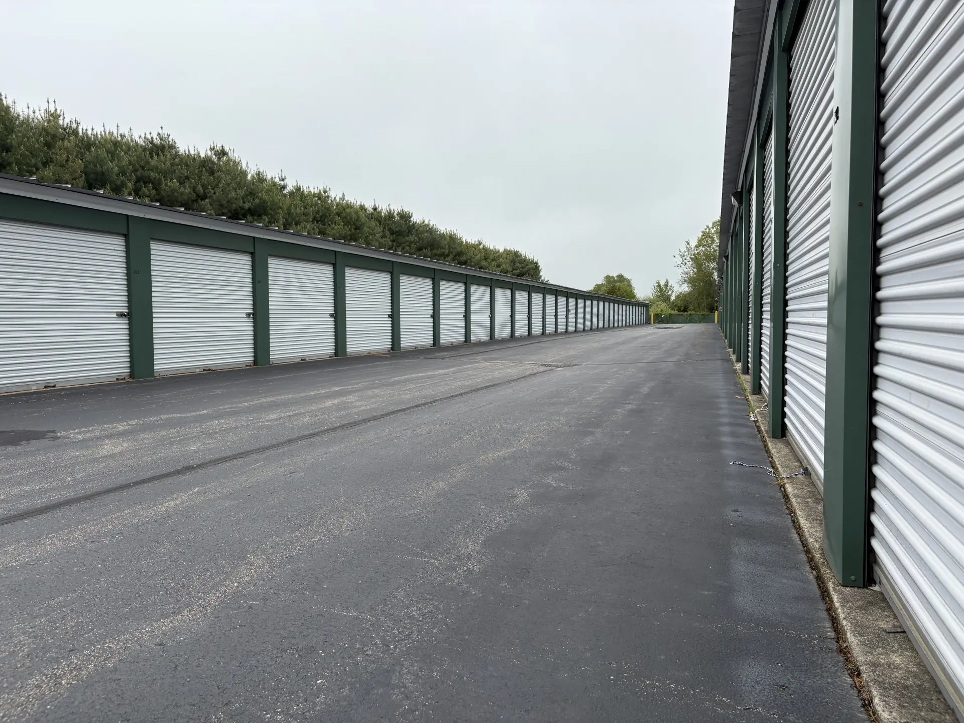 Outdoor row of storage units with white roll-up doors on both sides of a paved drive.