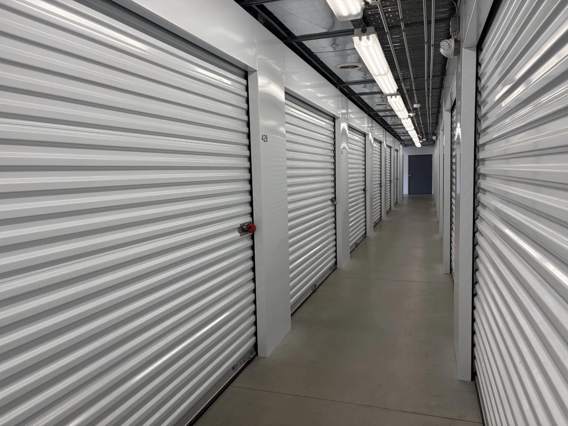 Interior hallway of storage facility with rows of white roll-up doors and overhead lights.