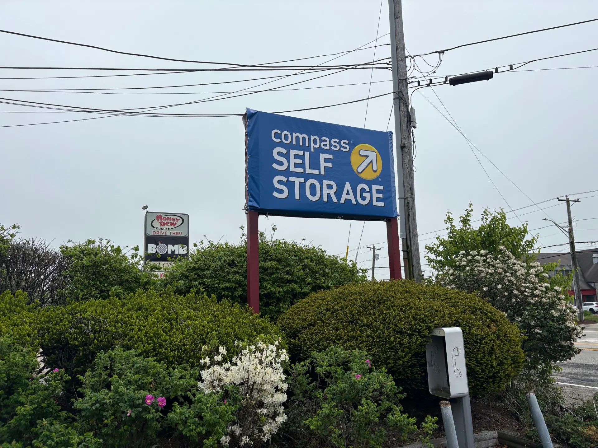 Compass Self Storage sign on a tall pole surrounded by shrubs, payphone, and power lines.