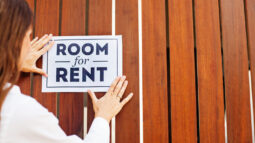 A woman placing a sign on a fence that says “Room for Rent.”