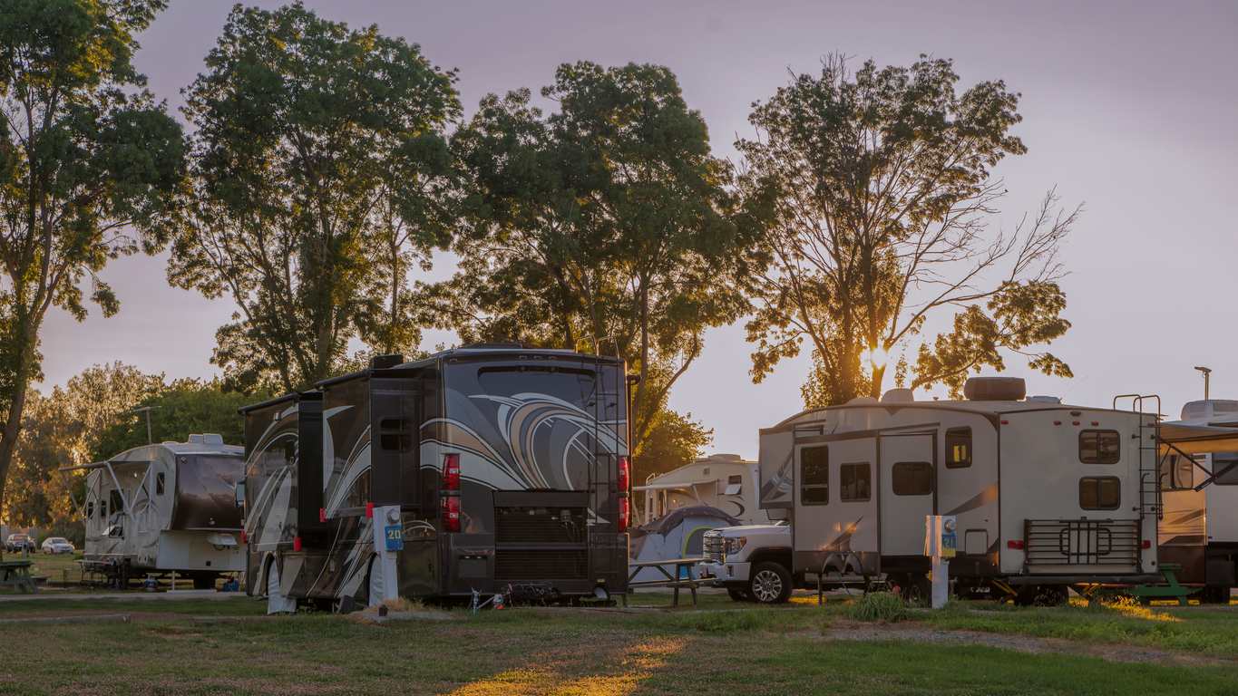 Several RVs are parked at a campground during sunset, with trees and warm evening light shining through.