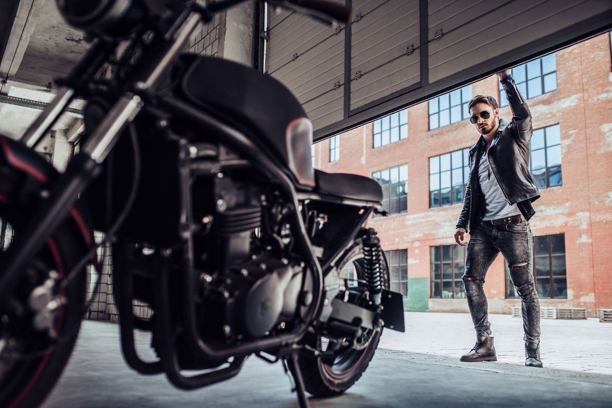 A man in a leather jacket and sunglasses raises a garage door with a black motorcycle parked inside.