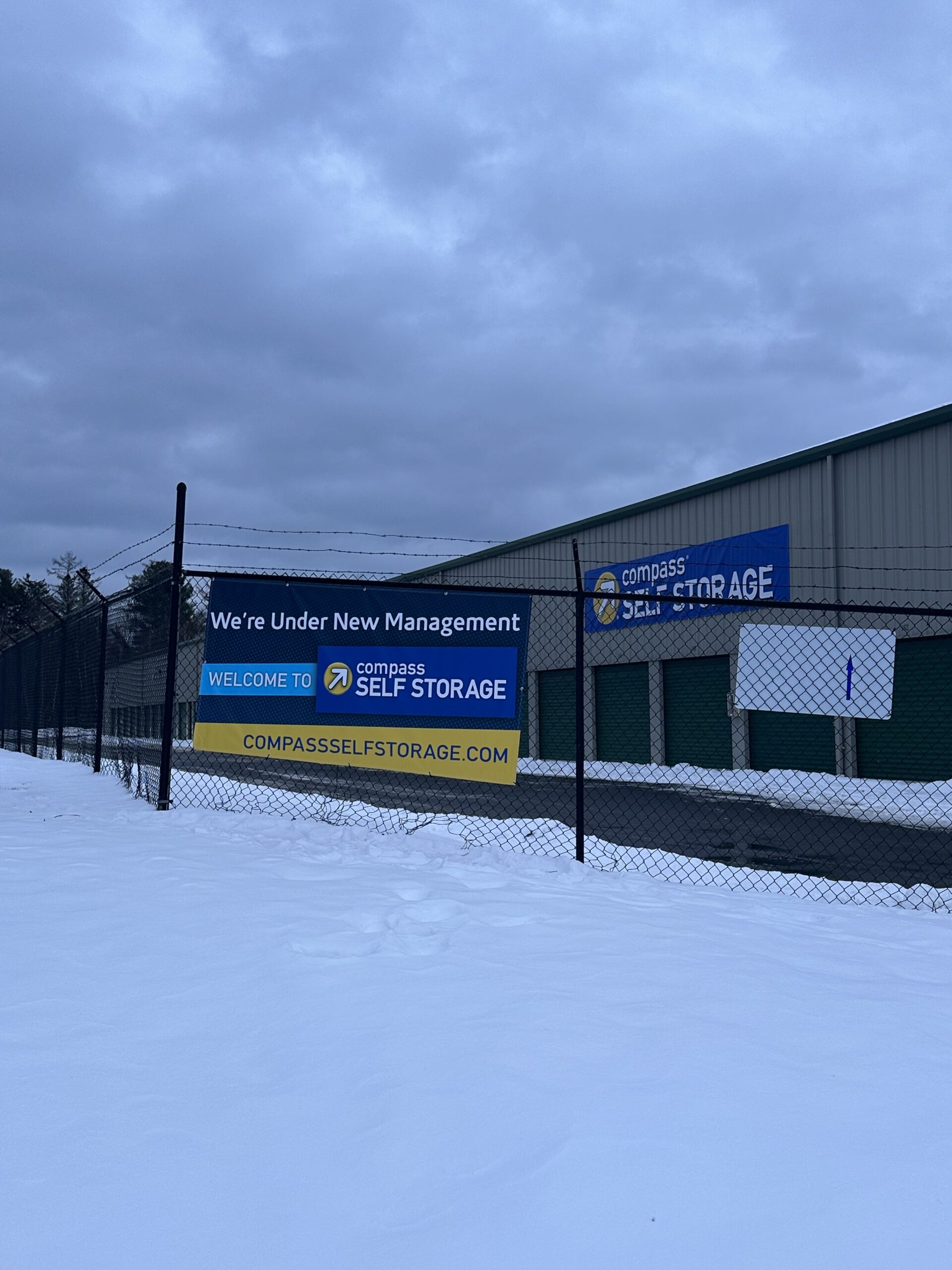 Snowy Compass Self Storage facility with 'New Management' banners, chain-link fence, and green storage units.