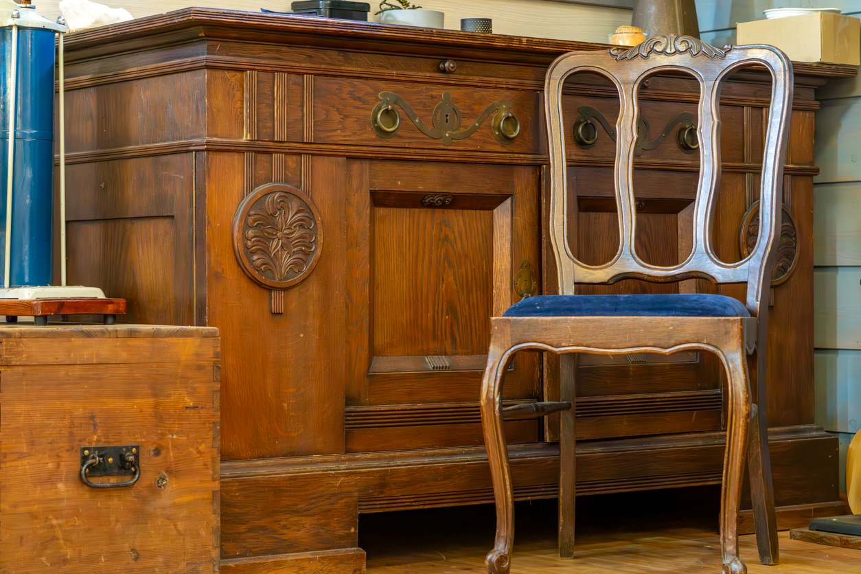 An old wooden dresser with carved details and a vintage wooden chair in a cozy room filled with old furniture pieces.