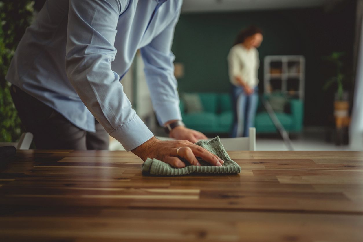A person wiping a wooden tabletop with a cloth while another person cleans in the living room.