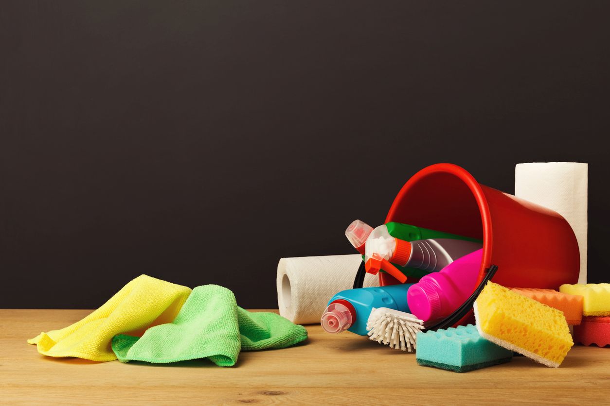 Cleaning supplies scattered on a wooden surface, including spray bottles, sponges, cloths, a red bucket, and towels.