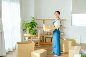 A young woman holding a cardboard box surrounded by moving boxes in a bright living room with wooden shelves, several chairs, and a plant.
