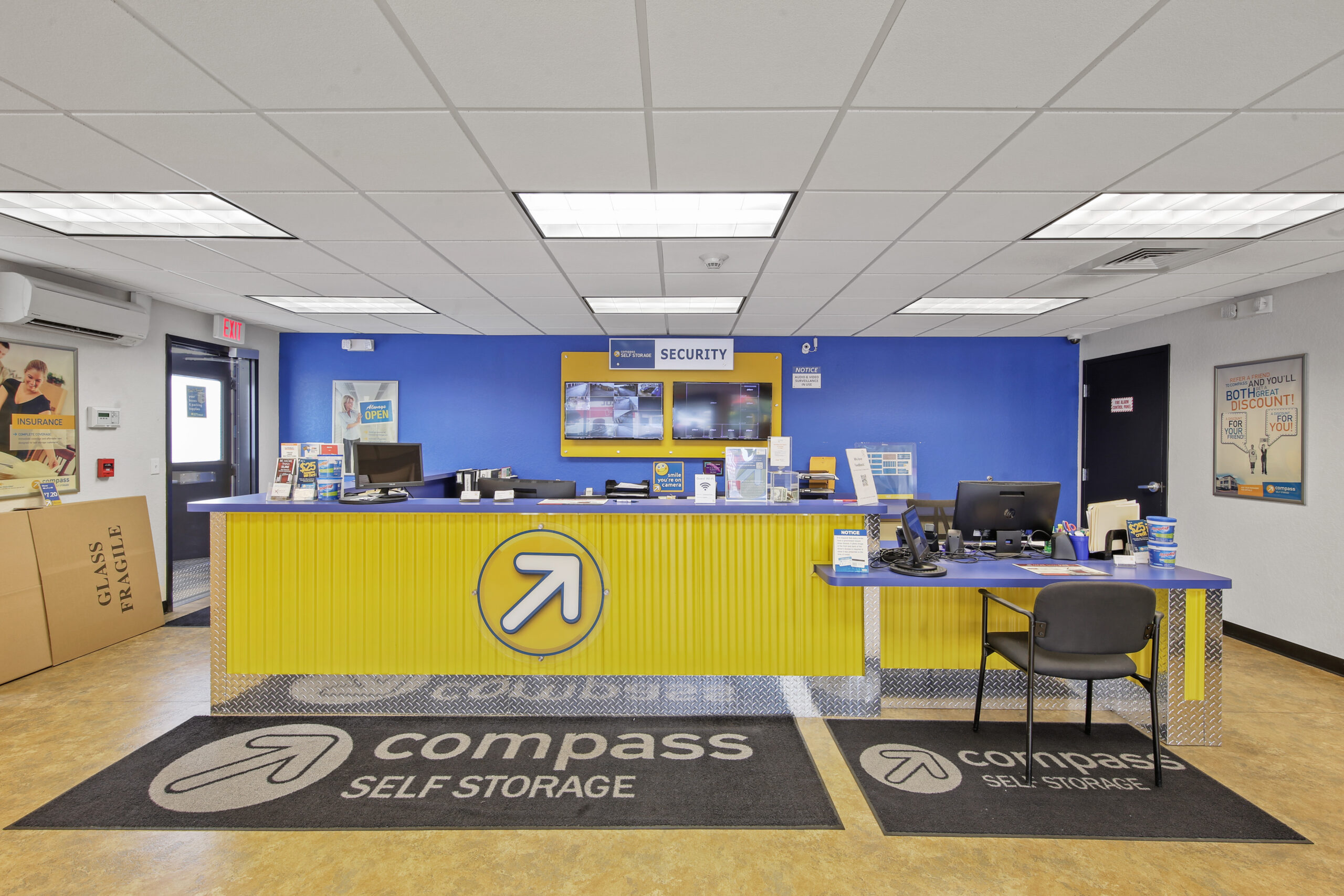 Compass Self Storage front desk with yellow and blue branding, security monitors, computers, and moving supplies.