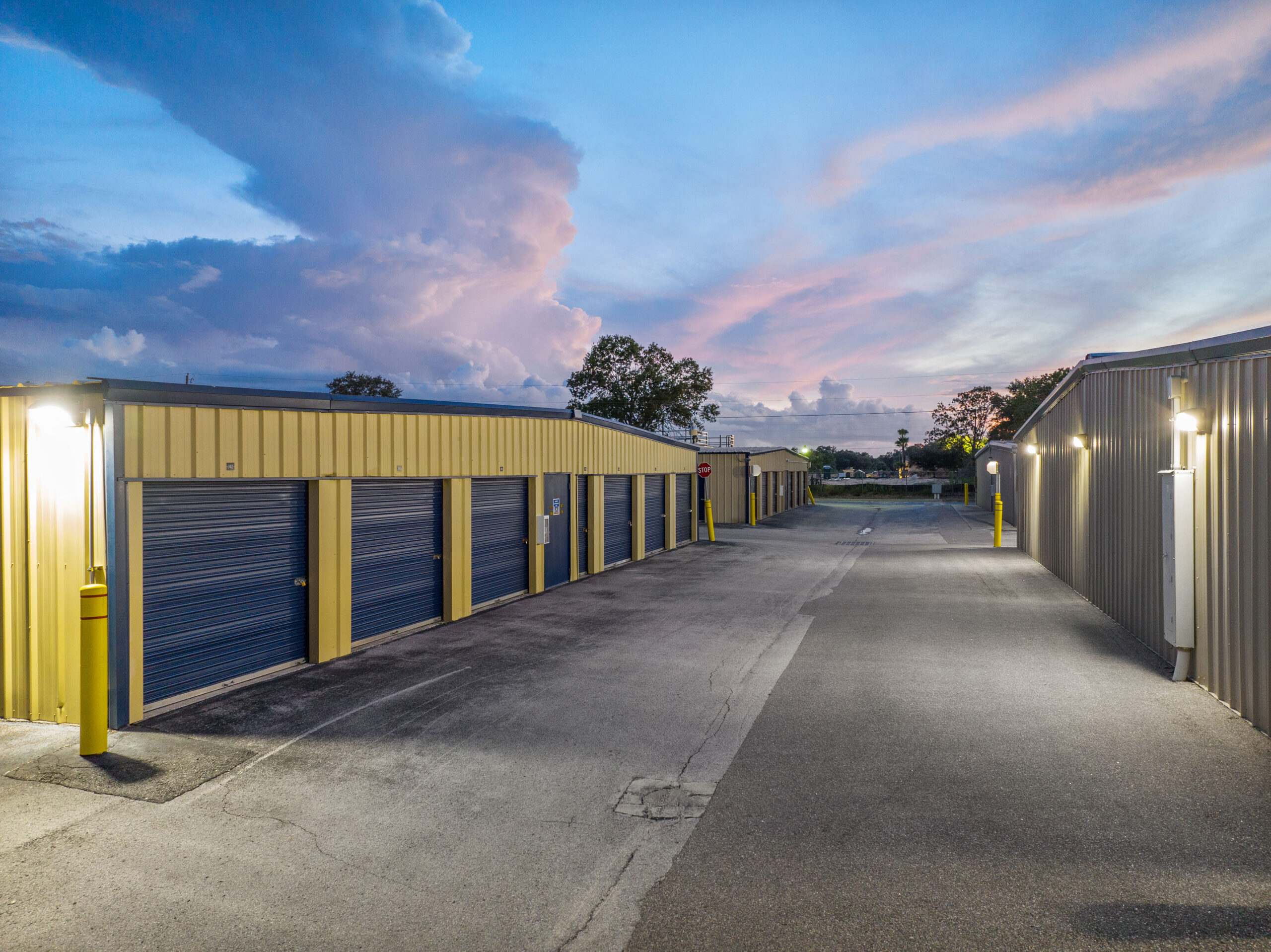 Evening view of self storage units with yellow siding, blue roll-up doors, and lighted pathways under a colorful sky.